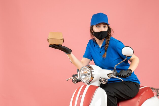 Vue de dessus d'un livreur choqué portant un masque médical et des gants assis sur un scooter livrant des commandes sur fond de pêche pastel