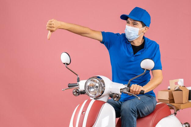 Vue de dessus d'un livreur nerveux en masque médical portant un chapeau assis sur un scooter et faisant un mauvais geste sur fond de pêche pastel