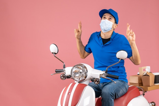 Vue de dessus d'un livreur plein d'espoir en masque médical portant un chapeau assis sur un scooter et rêvant de quelque chose sur fond de pêche pastel