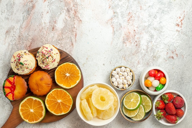 Vue de dessus de loin agrumes sur la planche bols de fraises limes bonbons et ananas séchés et tranches d'orange et de biscuits sur la planche à découper en bois