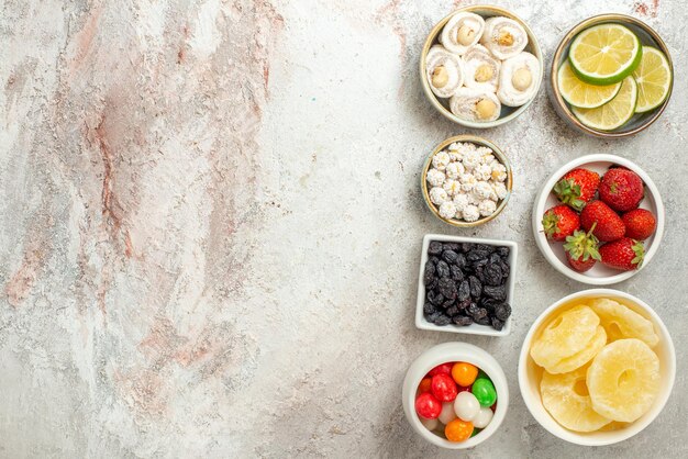 Vue de dessus de loin des bonbons dans des bols bols de différents bonbons et ananas séchés sur le côté droit de la table blanche