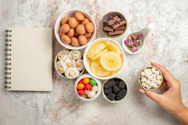 Vue de dessus de loin bonbons dans des bols cahier blanc à côté des bols de bonbons fruits secs et baies à la main