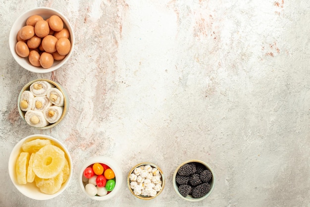 Vue de dessus de loin des bonbons dans des bols six bols de bonbons appétissants et d'ananas séchés sur fond blanc