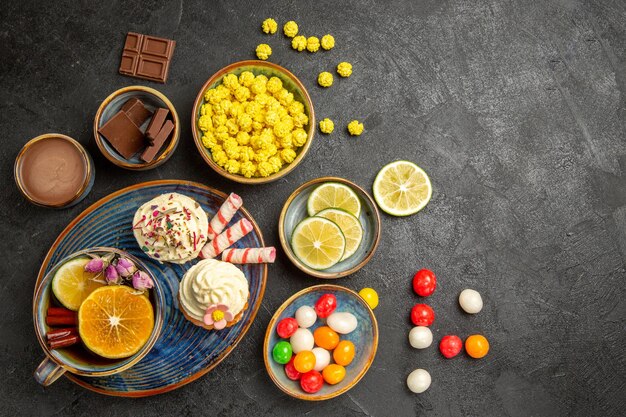 Vue de dessus de loin des bonbons sur la table des cupcakes et une tasse de thé au citron sur la soucoupe des bols de chocolat au citron et à la crème de chocolat des bonbons colorés sur la table sombre