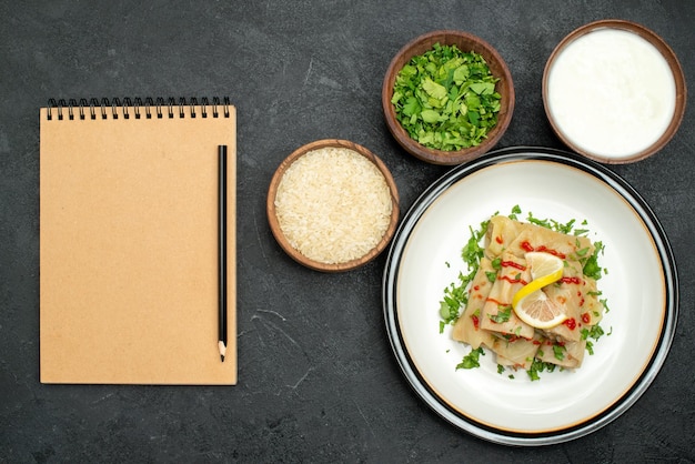 Vue de dessus de loin chou farci appétissant chou farci aux herbes citron et sauce sur assiette blanche et bols aux herbes de riz et crème sure à côté d'un carnet de crème avec un crayon sur un tableau noir