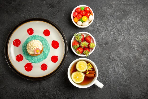 Vue de dessus de loin cupcake une tasse de thé assiette blanche de cupcake bols de fraises et de bonbons à côté de la tasse de thé avec des bâtons de cannelle et du citron