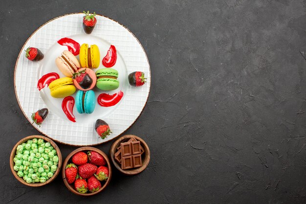 Vue de dessus de loin fraises et macarons assiette de fraises appétissantes sauce macarons français à côté des bols de bonbons sur la table sombre