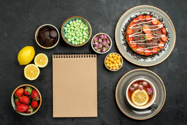 Vue de dessus de loin gâteau au thé gâteau appétissant thé noir citrons chocolat et différents bonbons à côté du cahier crème sur tableau noir