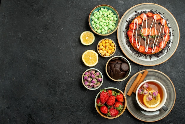 Vue de dessus de loin gâteau avec des bonbons gâteau avec du thé noir aux fraises avec des noisettes au citron des bols de chocolat et différents bonbons sur le côté droit de la table sombre