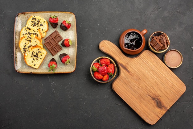 Vue de dessus de loin gâteau dessert avec fraises enrobées de chocolat et chocolat et planche à découper entre des bols de crème au chocolat et des baies sur la table