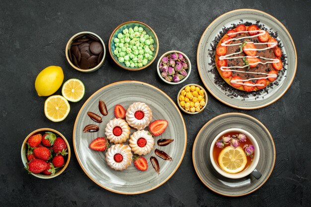 Vue de dessus de loin gâteau avec thé appétissant gâteau biscuits au thé noir avec fraise dans une assiette blanche à côté de citrons chocolat et différents bonbons sur tableau noir