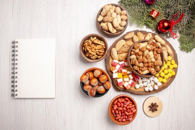 Vue de dessus de loin noix sur le plateau branches d'épicéa avec différents bonbons et cacahuètes sur le plateau de la cuisine à côté du cahier blanc bols de noisettes noix sur la table