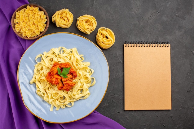 Vue de dessus de loin des pâtes et des bols de viande de pâtes et une assiette de sauce pour pâtes et de viande sur la nappe violette à côté du cahier crème sur fond sombre