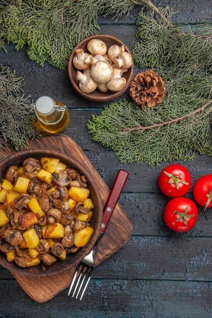 Vue de dessus de loin plat et branches plat de pommes de terre et champignons sur la planche à découper à côté de la fourchette et trois tomates sous bol d'huile de champignons blancs et branches d'épinette