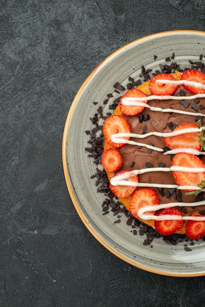 Vue de dessus de loin tarte au dessert appétissante avec fraise et chocolat sur une assiette blanche sur le côté gauche de la table sombre