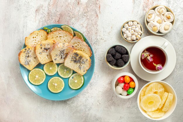 Vue de dessus de loin tasse de thé assiette de gâteau et citrons verts tranchés à côté des bols de différents bonbons ananas séchés Délice turc et tasse de thé sur fond blanc