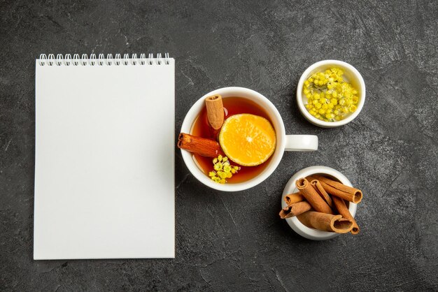 Vue de dessus de loin une tasse de thé avec un cahier blanc au citron à côté de la tasse de thé au citron et des bols de baies et de bâtons de cinabre au centre de la table