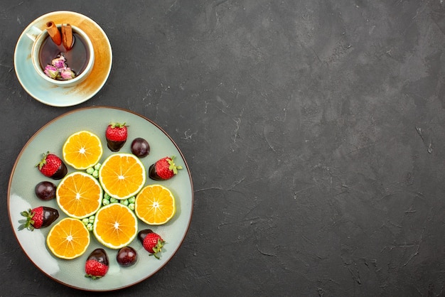 Vue de dessus de loin thé avec des fruits fraises enrobées de chocolat appétissants bonbons oranges et verts hachés à côté d'une tasse de thé avec des bâtons de cannelle sur le côté gauche de la table sombre