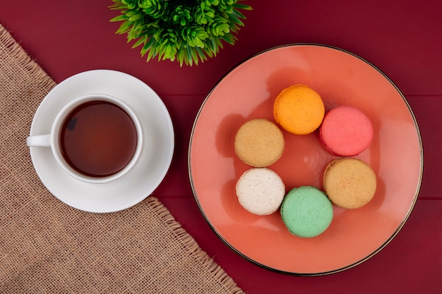 Vue de dessus des macarons colorés sur une assiette avec une tasse de thé sur une surface rouge