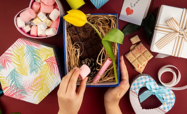 Vue de dessus de la main féminine mettant la guimauve dans une boîte actuelle avec une fleur de tulipe de couleur jaune, une barre de chocolat noir, un cône et de la paille sur un tableau rouge foncé avec une boîte remplie de guimauve