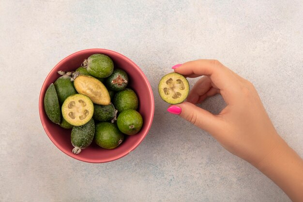 Vue de dessus de la main féminine tenant une douce moitié de feijoa avec feijoas sur un bol sur un mur gris