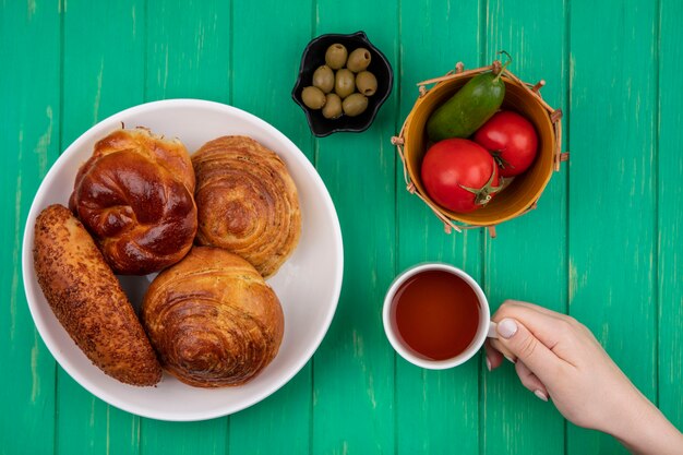 Vue de dessus de la main féminine tenant une tasse de thé avec des petits pains sur une plaque blanche avec des olives sur un bol noir avec des tomates et des concombres sur un seau sur un fond vert