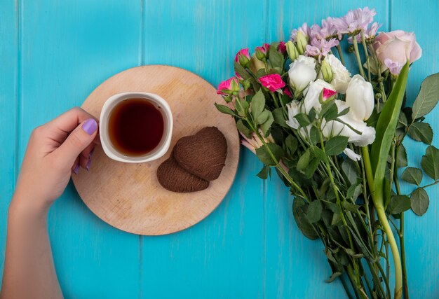 Vue de dessus de la main féminine tenant une tasse de thé sur une planche de cuisine en bois avec de merveilleuses fleurs fraîches isolées sur un fond en bois bleu