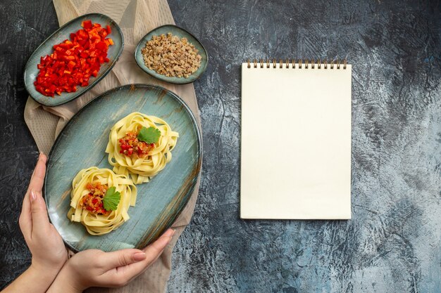 Vue de dessus de la main tenant une assiette bleue avec un délicieux repas de pâtes servi avec de la tomate et de la viande pour le dîner sur une serviette de couleur beige ses ingrédients à côté d'un cahier à spirale