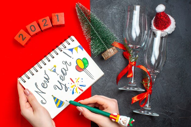 Vue de dessus de la main tenant le cahier à spirale avec dessin de nouvel an et numéros de gobelets en verre de sapin de Noël sur fond sombre et rouge