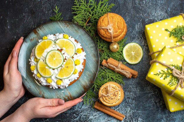 Vue de dessus d'une main tenant un délicieux gâteau crémeux et des branches de sapin citron cannelle limes jaunes coffrets cadeaux sur fond sombre