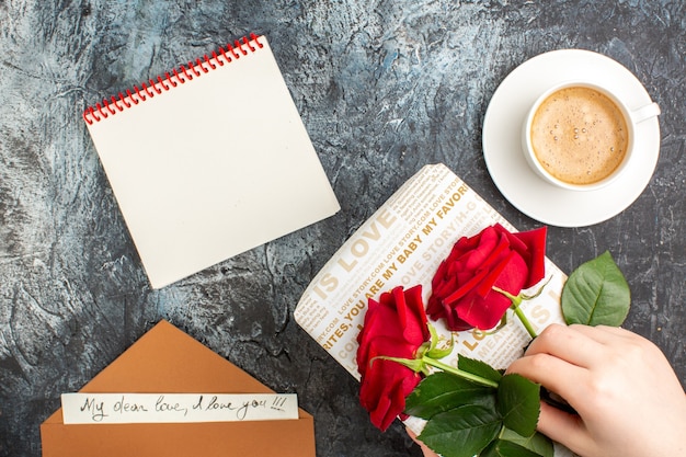 Vue de dessus de la main tenant des roses rouges sur une belle boîte-cadeau et une tasse d'enveloppe de café avec un cahier de lettre d'amour sur une surface sombre et glacée