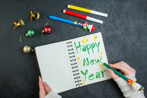 Vue de dessus de la main tenant un stylo sur un cahier à spirale avec des accessoires de décoration d'écriture de bonne année sur fond noir