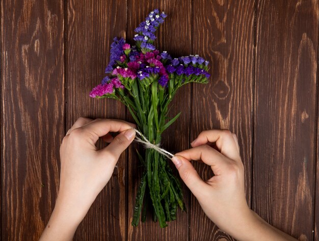 Vue de dessus des mains attachant avec une corde un bouquet de fleurs de limonium statice couleur rose et violet sur fond rustique en bois