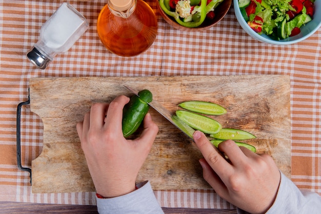 Vue de dessus des mains couper le concombre avec un couteau sur une planche à découper avec salade de légumes sel de beurre fondu sur la surface du tissu à carreaux