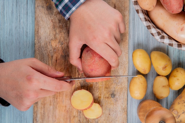 Vue de dessus des mains couper la pomme de terre avec un couteau sur une planche à découper avec d'autres dans le panier et sur la surface en bois