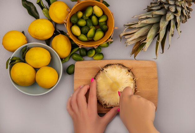 Vue de dessus des mains féminines ananas de coupe sur une planche de cuisine en bois avec un couteau avec des citrons sur un bol avec des kinkans sur un seau sur une surface blanche