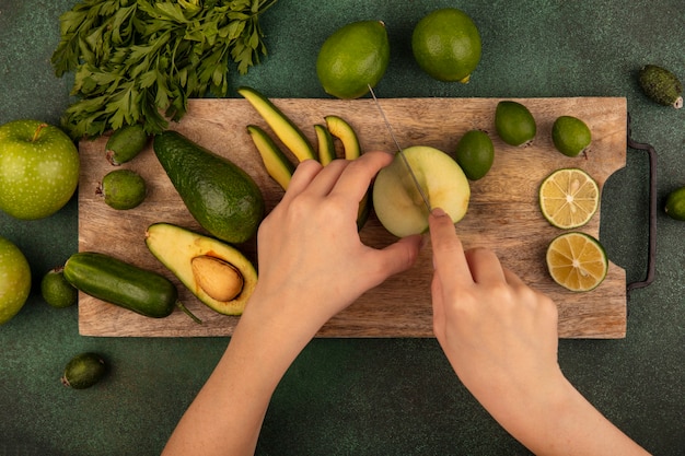 Vue de dessus des mains féminines coupant la pomme fraîche avec un couteau sur une planche de cuisine en bois avec limes feijoas avocats pommes vertes et persil isolé sur un mur vert