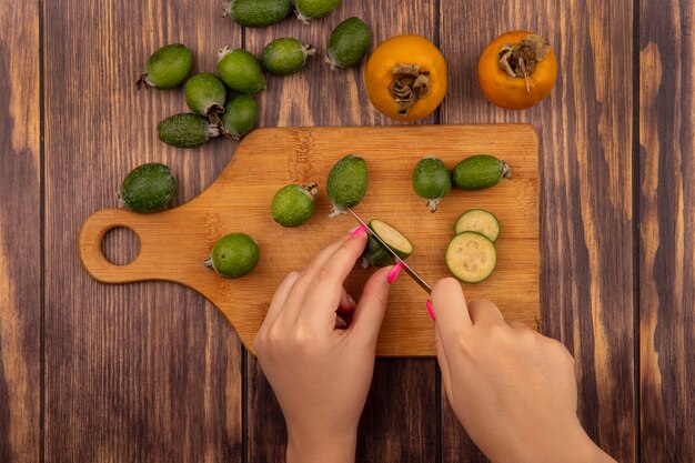 Vue de dessus des mains féminines coupe feijoa sur une planche de cuisine en bois avec un couteau avec des fruits de kaki frais et feijoas isolés sur un mur en bois