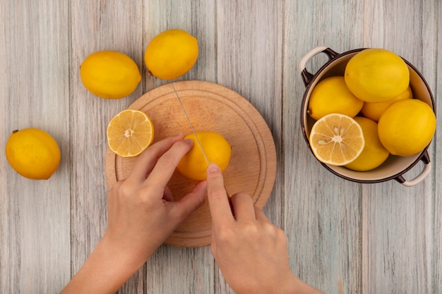 Vue de dessus des mains féminines couper le citron sur une planche de cuisine en bois avec un couteau avec des citrons sur un bol avec des citrons isolé sur une surface en bois gris