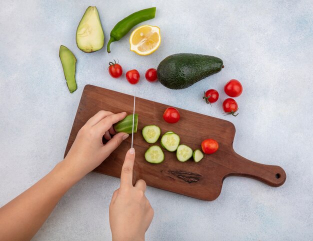 Vue de dessus des mains féminines couper le concombre en tranches sur planche de cuisine avec couteau avec avocat tomates cerises citron isolé sur blanc