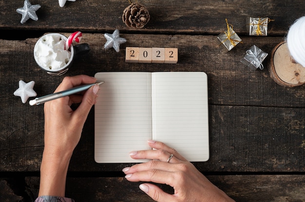 Vue de dessus des mains féminines écrivant une résolution du nouvel an 2022 et des plans dans un cahier sur un bureau en bois décoré de vacances