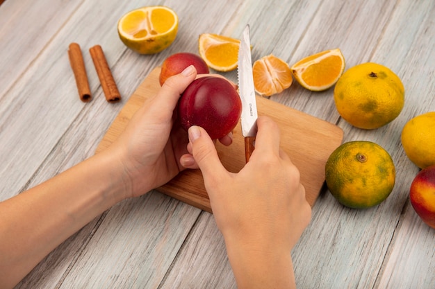 Vue de dessus des mains féminines hacher une pêche sur une planche de cuisine en bois avec un couteau avec des mandarines isolé sur un fond en bois gris