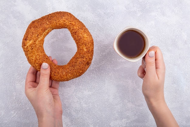 Vue de dessus des mains féminines tenant un bagel turc au sésame avec une tasse de thé sur fond blanc