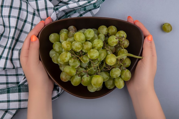 Vue de dessus des mains féminines tenant bol de raisin blanc sur tissu à carreaux et fond gris