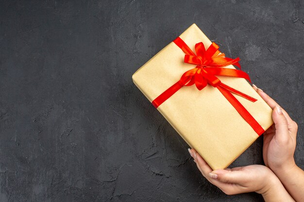 Vue de dessus des mains féminines tenant un cadeau de Noël dans du papier brun attaché avec un ruban rouge sur une surface sombre