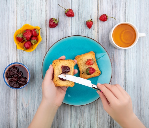 Vue de dessus des mains féminines tenant du pain grillé avec de la confiture de fraises sur une plaque bleue avec des fraises fraîches sur un bol jaune sur un fond en bois gris