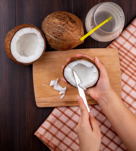 Vue de dessus des mains féminines tenant la noix de coco dans une main et la coupe dans l'autre main sur une planche de cuisine en bois avec des noix de coco et un verre d'eau sur une nappe à carreaux sur fond noir