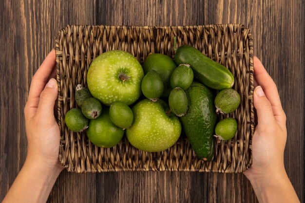 Vue de dessus des mains féminines tenant un plateau en osier de fruits frais tels que les pommes vertes feijoas limes sur une surface en bois