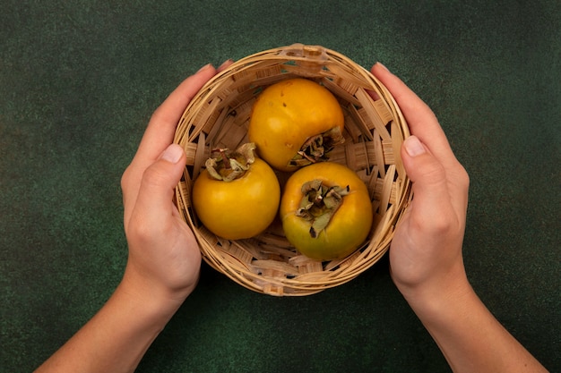 Vue de dessus des mains féminines tenant un seau de fruits kaki sur une surface verte