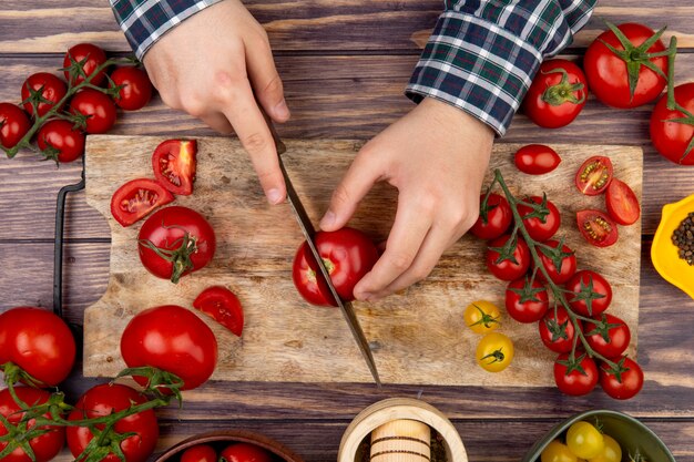 Vue de dessus des mains de femme coupant la tomate sur une planche à découper avec un couteau et un broyeur d'ail au poivre noir sur une surface en bois